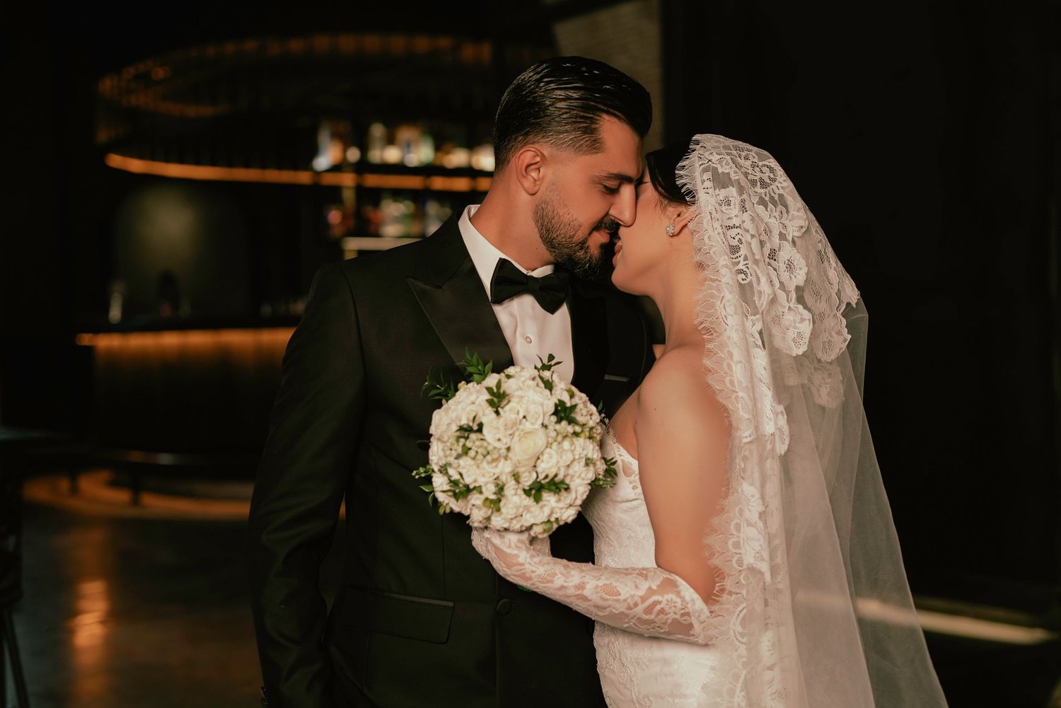 A romantic wedding couple shares a kiss in a dimly lit venue while the bride holds a white rose bouquet.