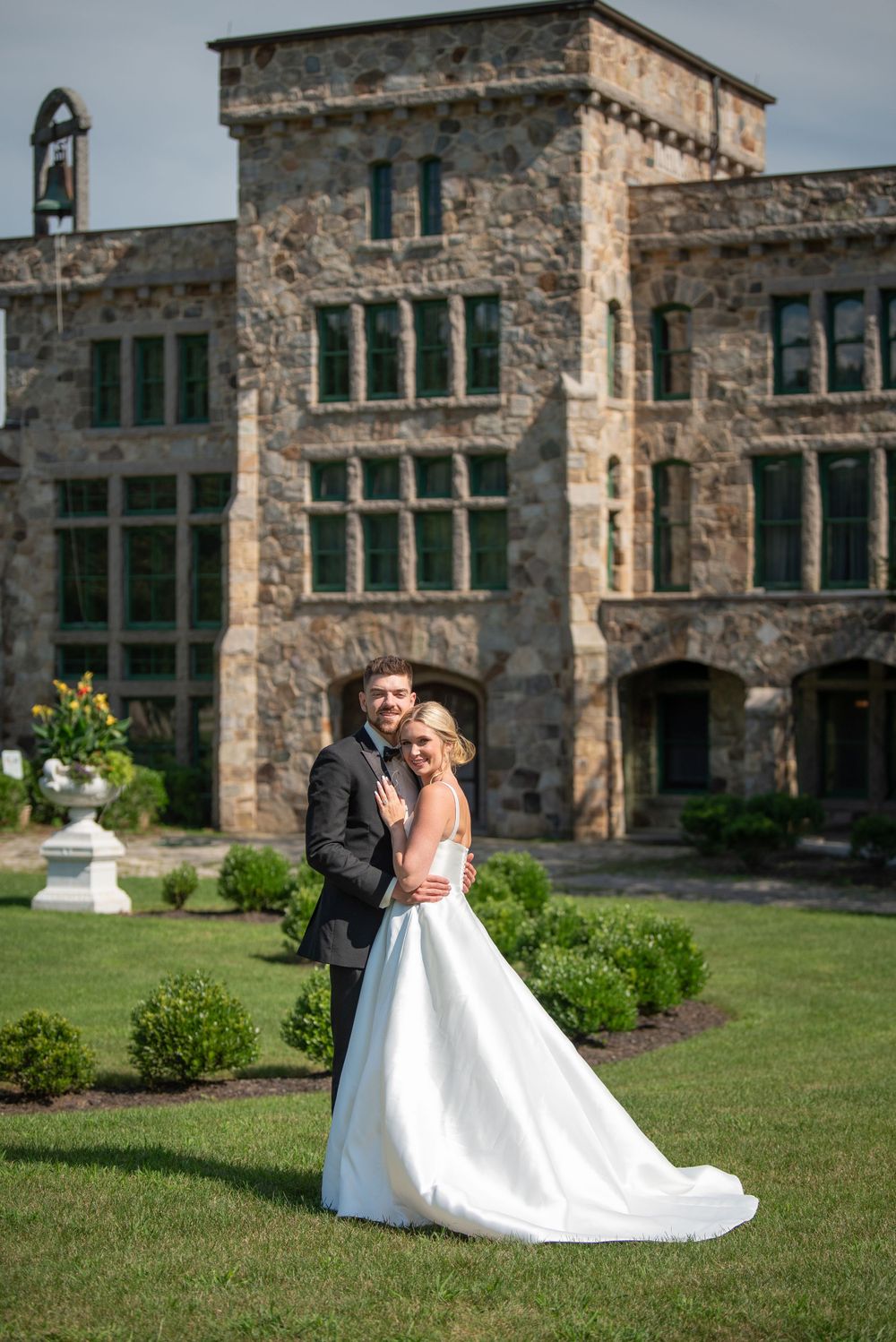 Wedding couple poses in front of a grand stone castle-like building with manicured grounds.