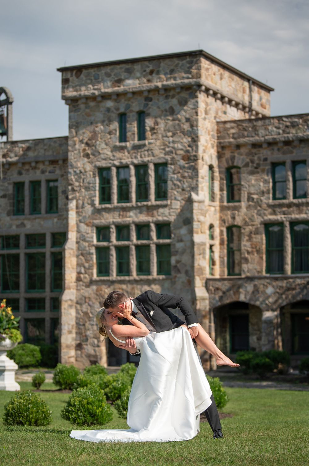 Couple performs playful dance move on lawn in front of stone castle-like building.