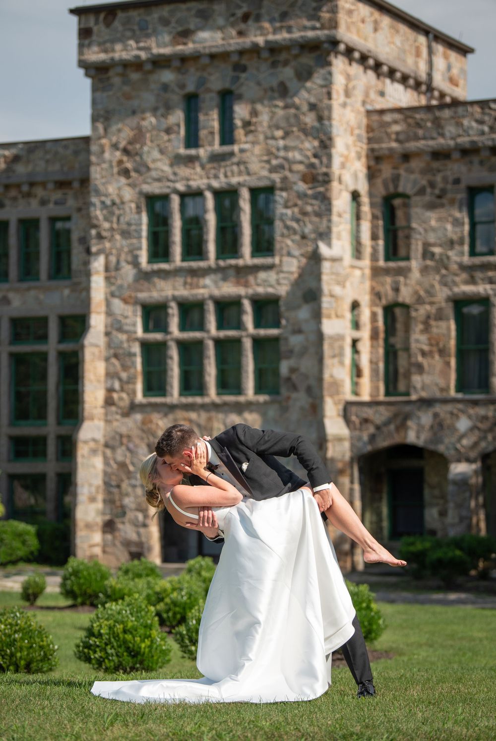 Wedding couple dips dramatically on grass with historic stone building backdrop.