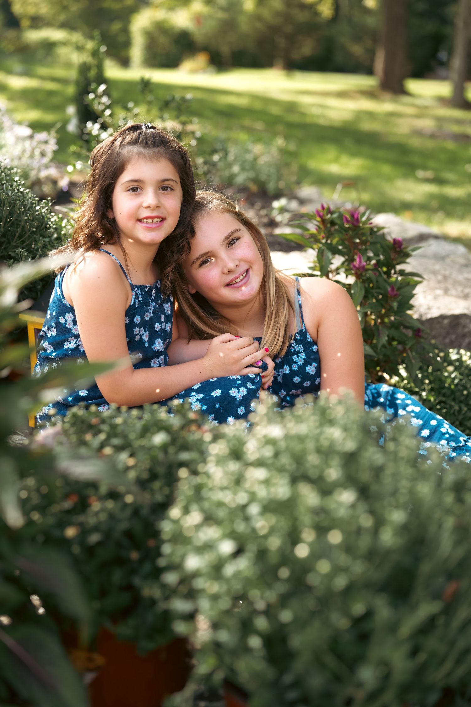 Two people in blue floral dresses posing together among garden flowers on a sunny day.
