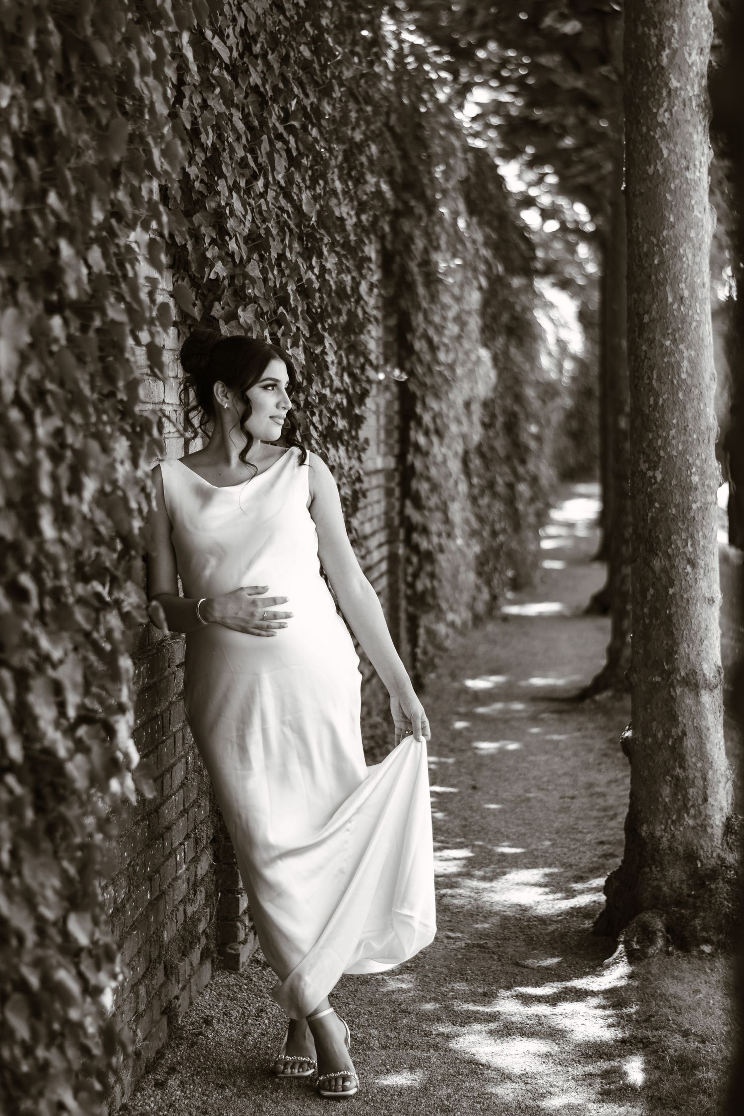 Artistic black and white photo of woman in flowing white dress walking through tree-lined path.