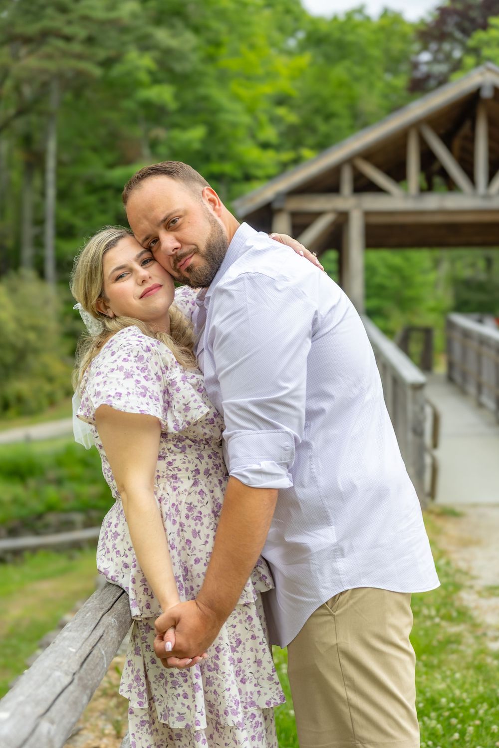 Couple embracing on a wooden bridge surrounded by lush green trees, woman in floral dress holding hands with man in white shirt.