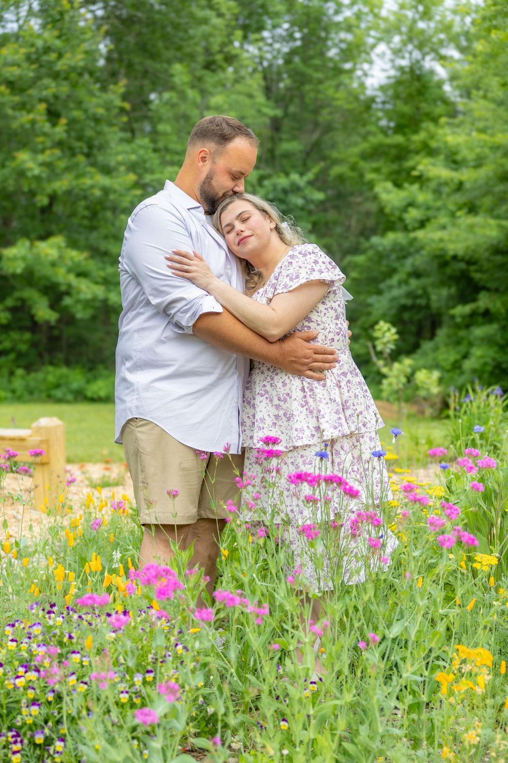 Couple embracing in a colorful wildflower garden, surrounded by pink and yellow blooms with lush green trees behind them.