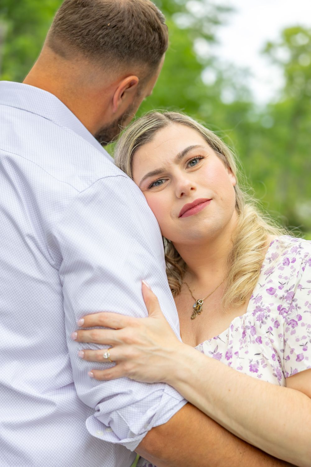 A woman in a floral dress rests her head on a man's shoulder while embracing outdoors among green trees.