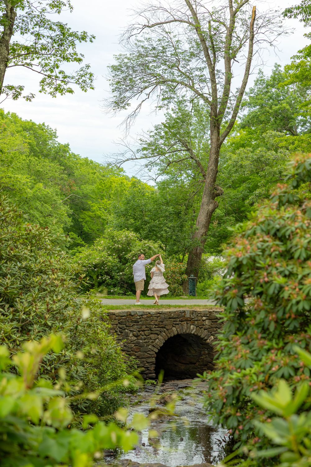 Couple dancing on a stone bridge surrounded by lush green trees and a serene stream in a romantic garden setting.
