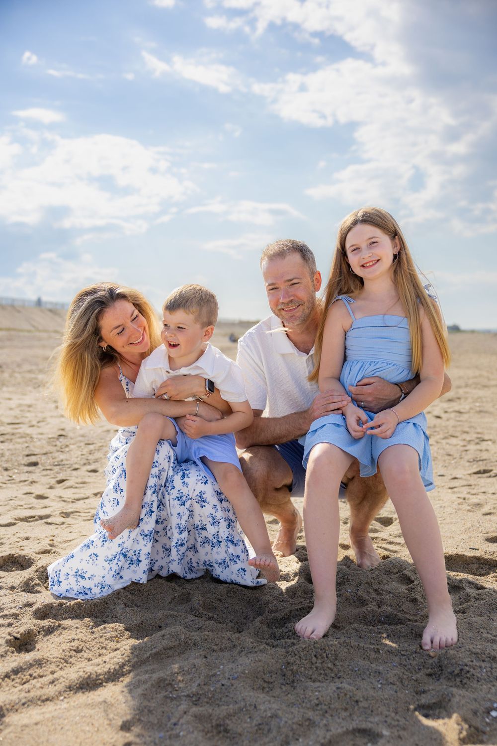 Happy family of four smiling together on a sandy beach on a sunny day, wearing blue and white outfits.