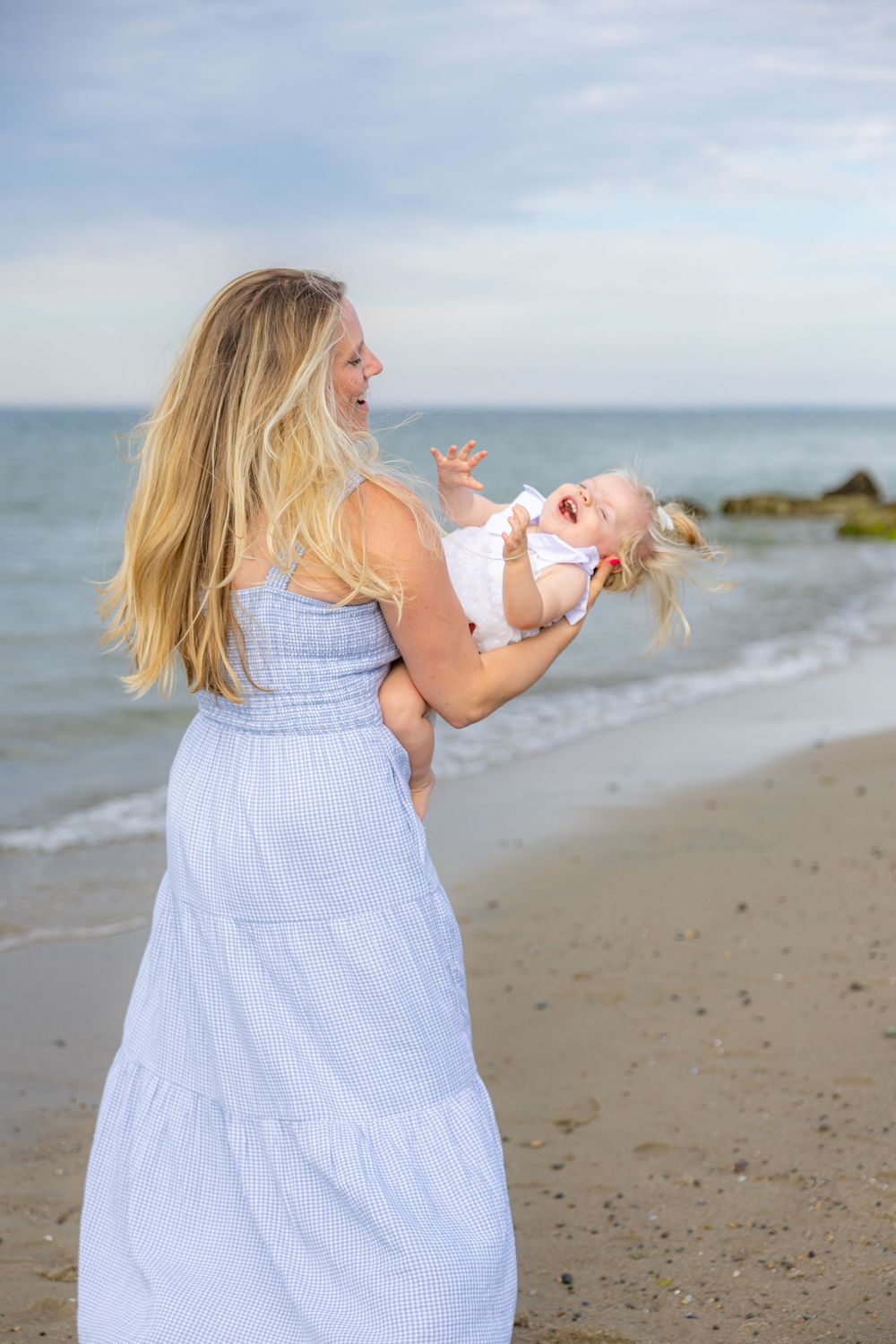 A joyful blonde mother in a white dress holds her laughing baby on a sunny beach with ocean waves behind them.
