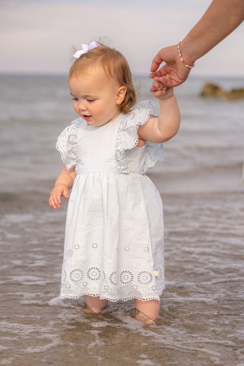 Toddler girl in white lace dress walking in ocean waves, holding an adult's hand at the beach.