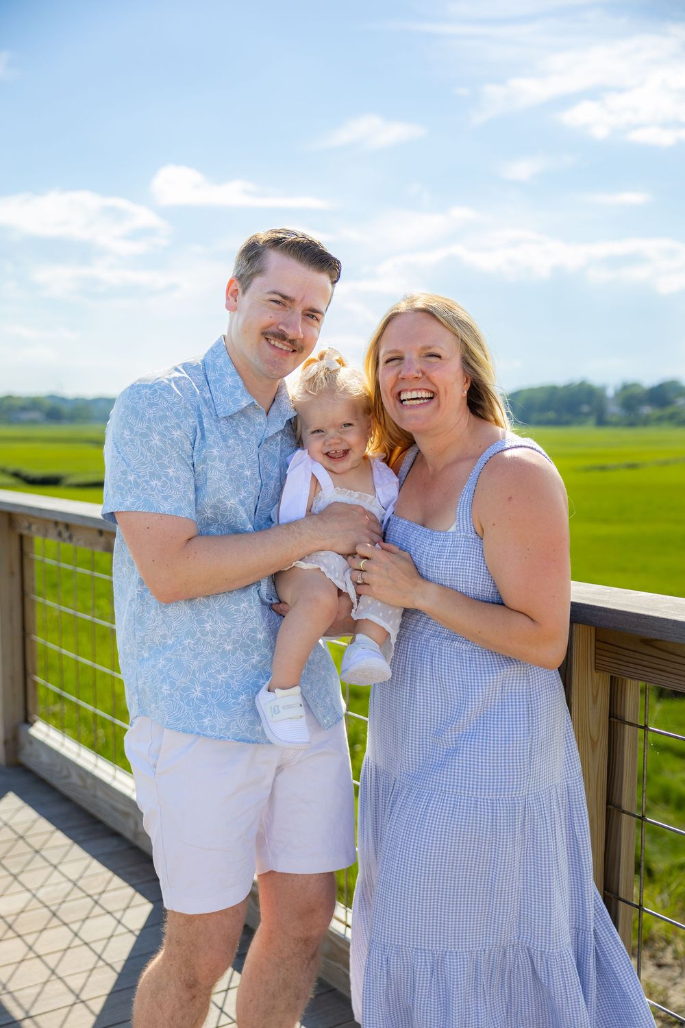 Happy family of three posing on a wooden bridge overlooking lush green marshlands on a sunny day.