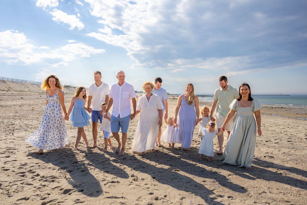 Large family group walking together on a sunny beach, dressed in coordinated pastel outfits, holding hands and smiling.