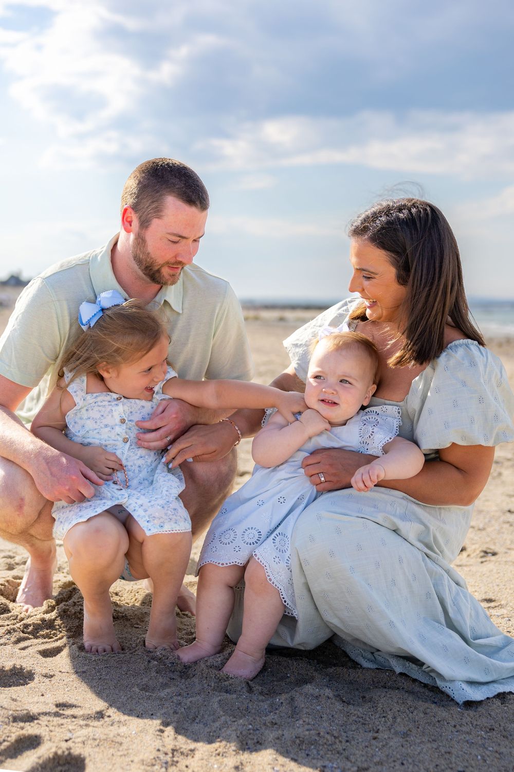 Family of four enjoying a sunny beach day, with parents smiling at their two young daughters sitting in the sand.