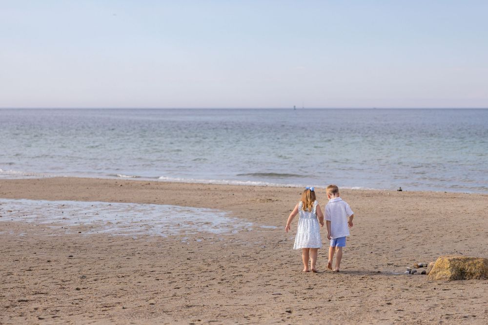 Two children walking hand in hand on a sandy beach toward the calm blue sea on a clear day.
