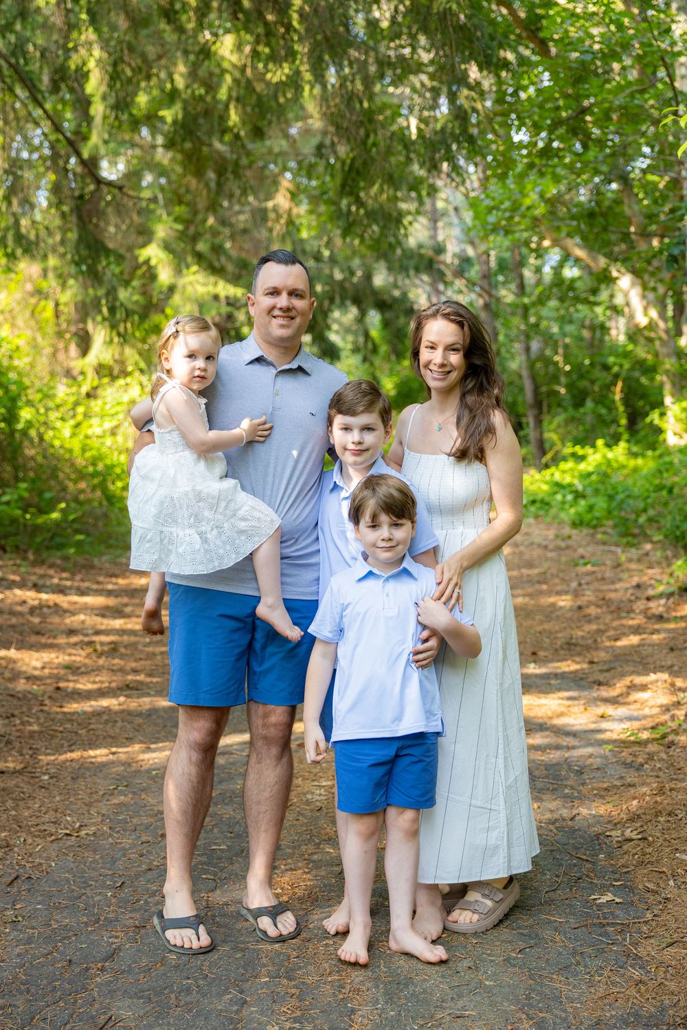 Family of five posing on a shaded garden path, parents with three young children wearing coordinated blue and white outfits.