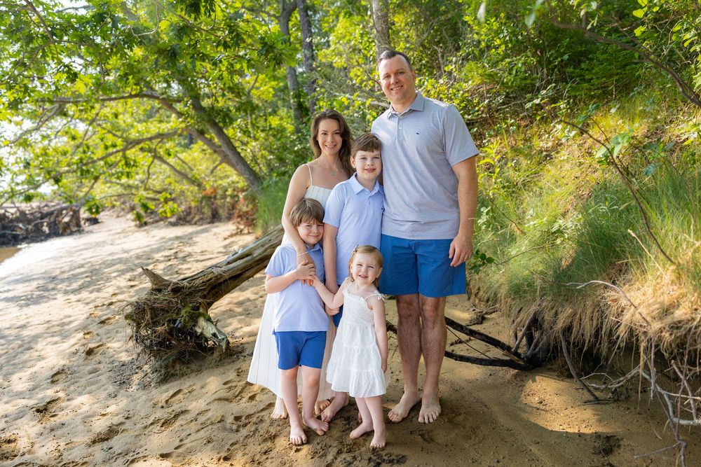 Family of five posing on sandy beach under lush green trees, parents with three young children in coordinating blue and white outfits.