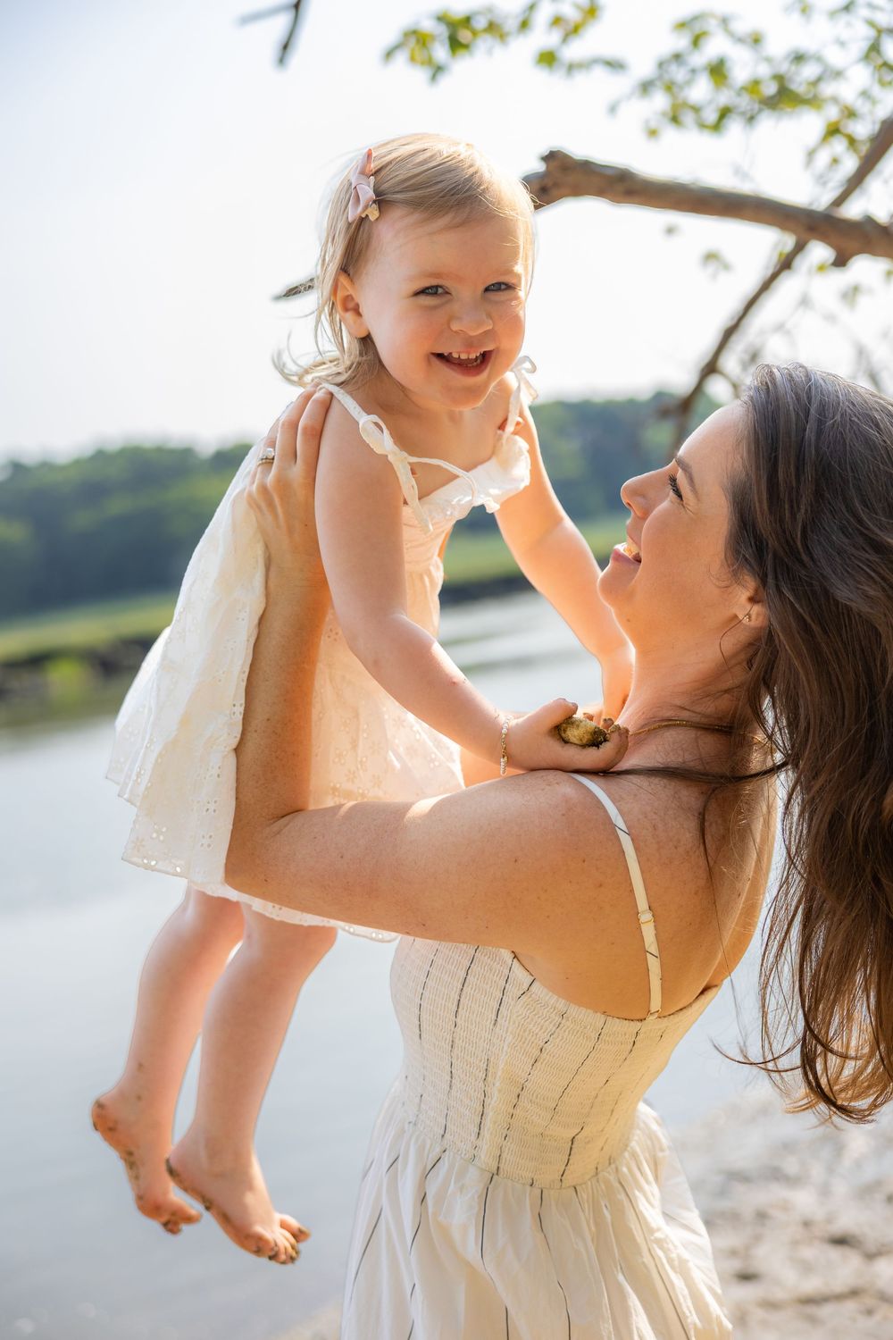 A joyful mother lifts her laughing toddler daughter outdoors near a sunlit lake, both wearing light summer dresses.