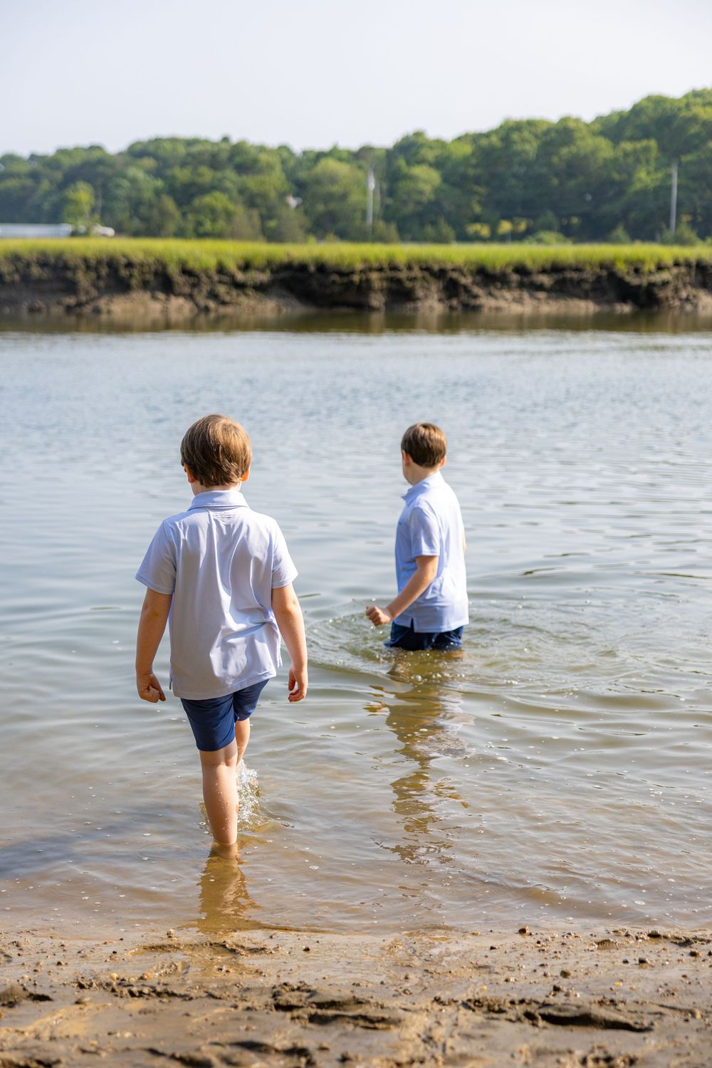 Two boys wading in shallow water near a sandy shore with green trees and grass in the background.