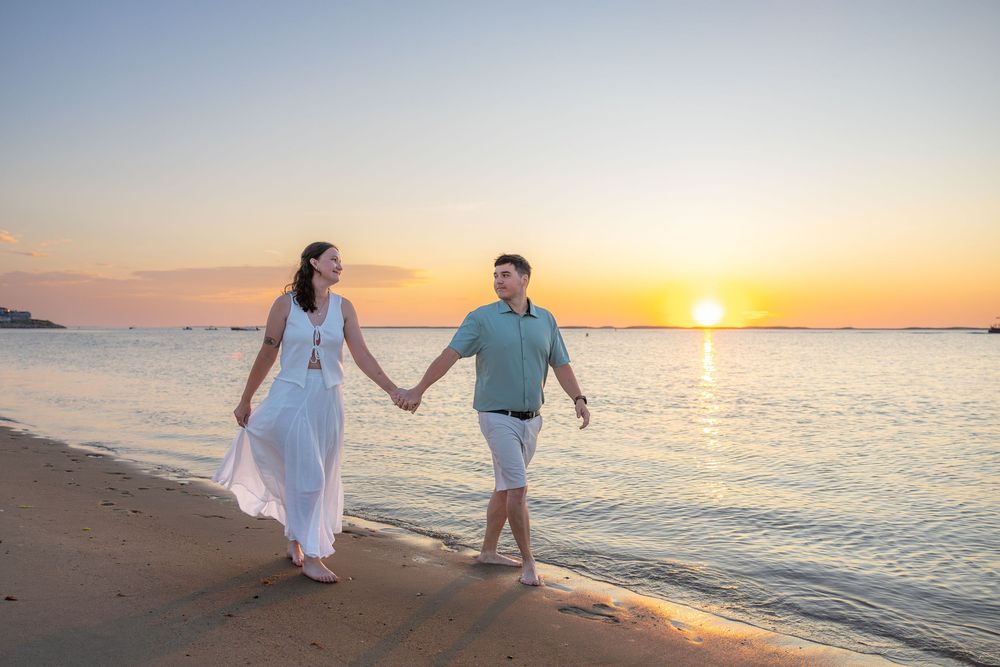 Couple holding hands walking on sandy beach at golden sunset with calm ocean waters in background.