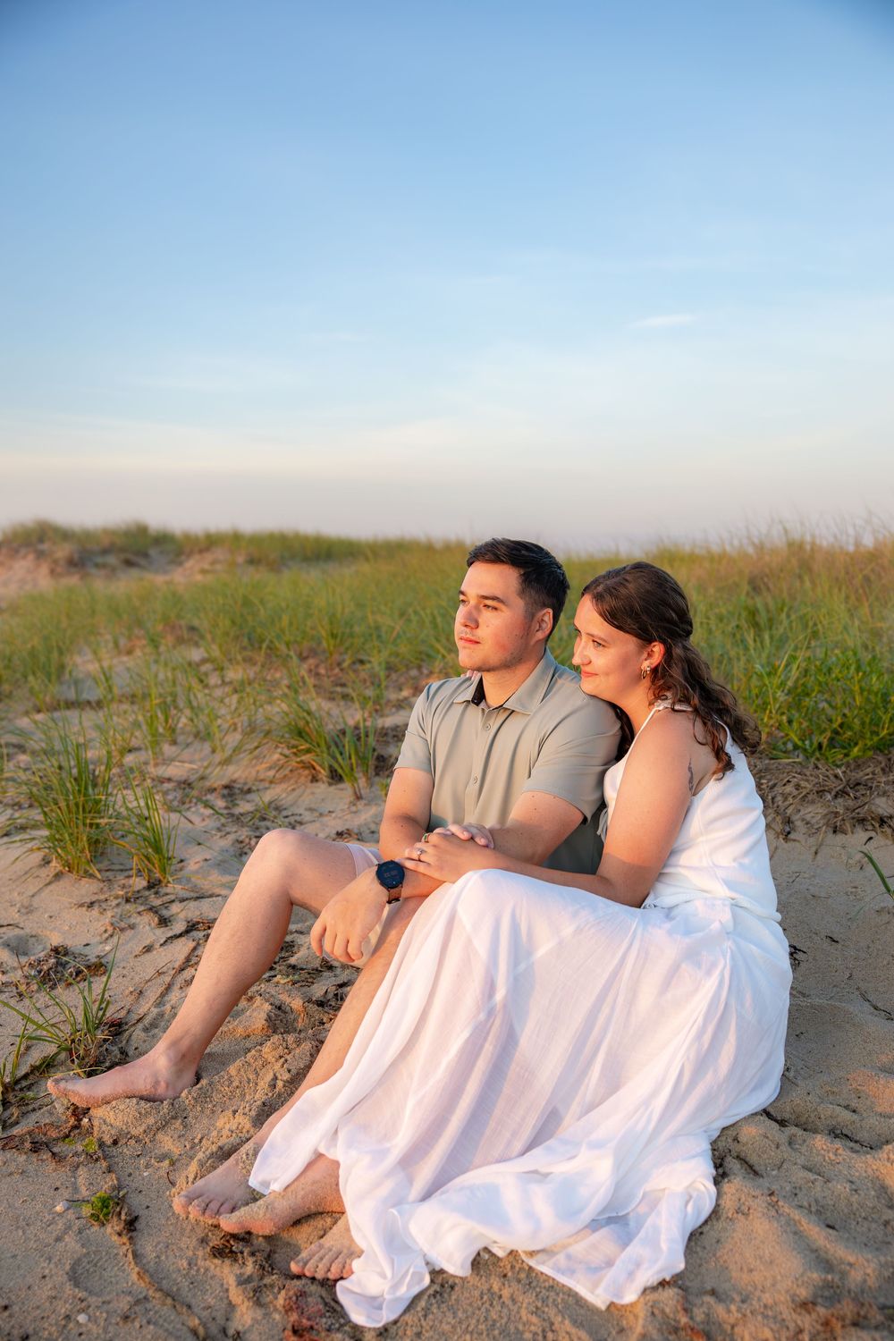 A couple sits together on sandy beach dunes at sunset, woman in white dress leaning on man in gray polo.
