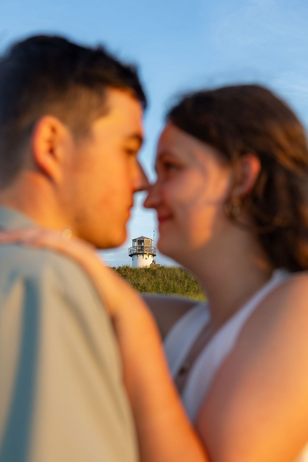 Couple sharing a romantic moment with a lighthouse on a grassy hill in the golden hour background.
