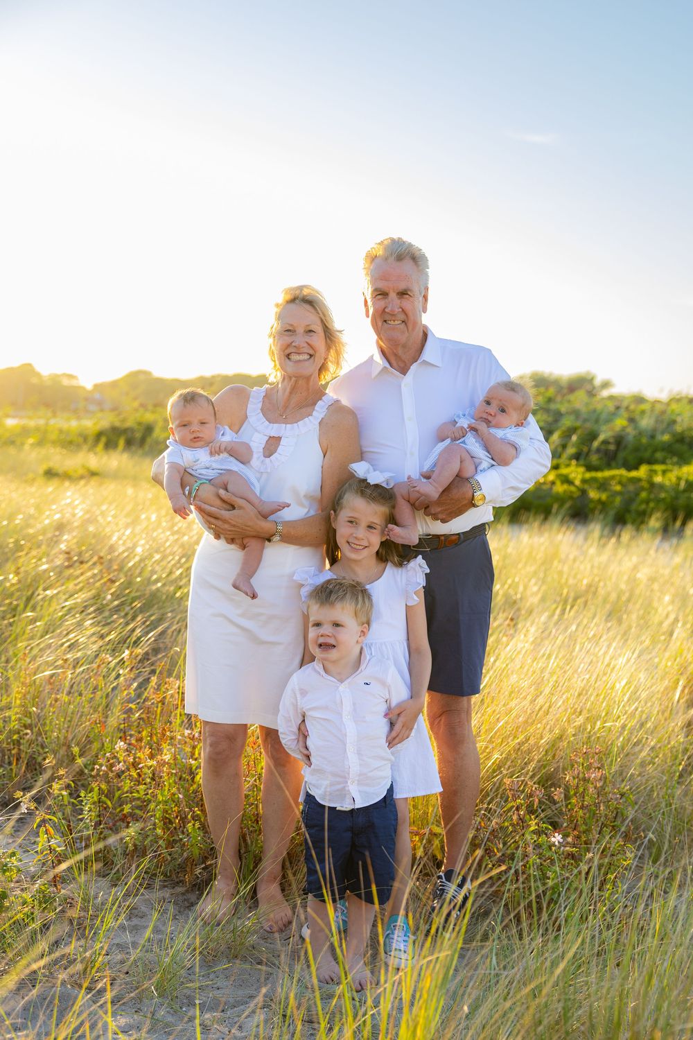 A family in white outfits poses together in a sunlit meadow of tall grass during golden hour.
