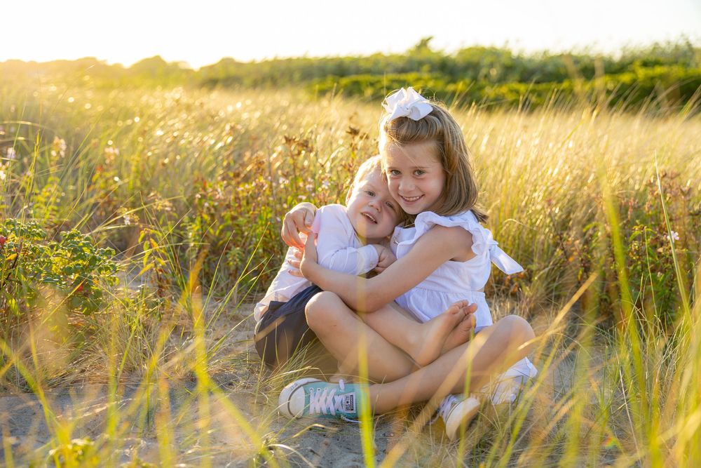 Two children sit together in a golden sunlit field of tall grass during a warm summer evening.