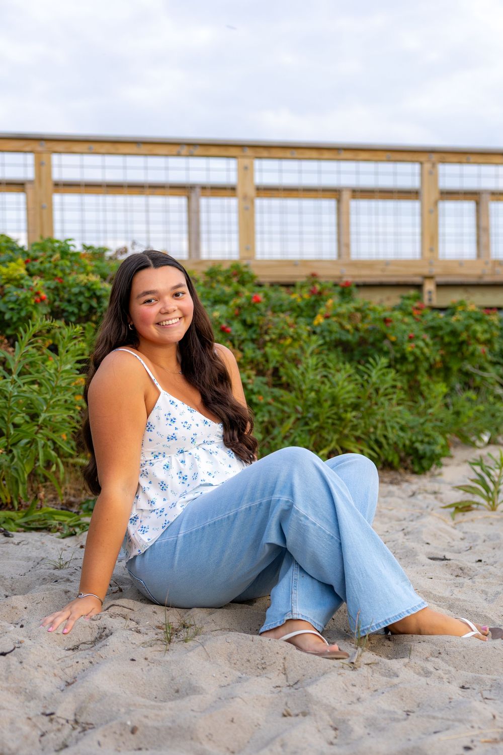 A person in a floral top and jeans poses casually on a sandy beach with a wooden boardwalk in the background.