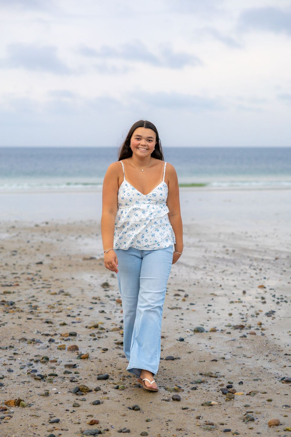 A person in light blue jeans and floral top poses casually on a rocky beach with ocean waves in the background.