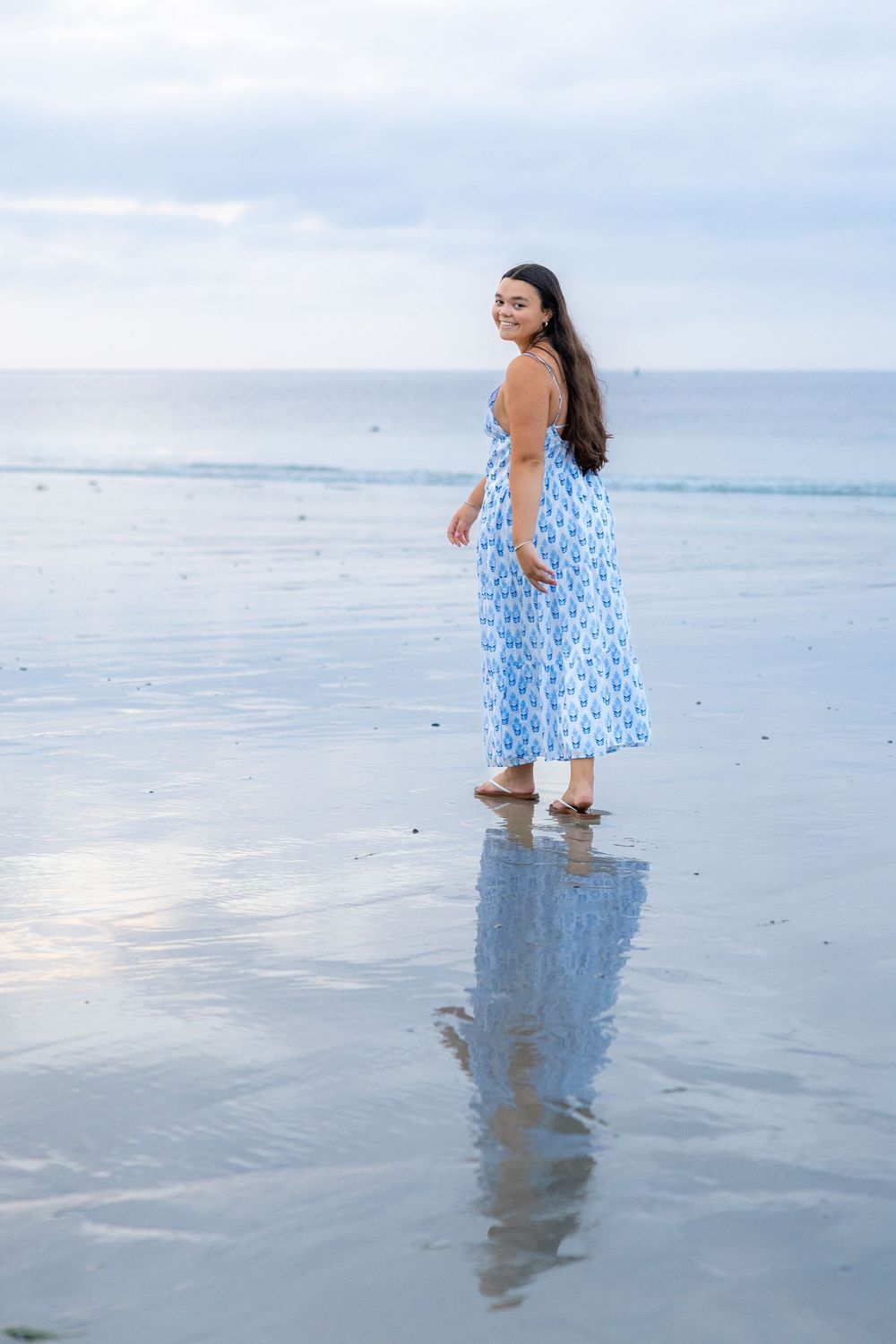 A person in a blue polka dot dress walks joyfully through shallow ocean water at the beach during sunset.
