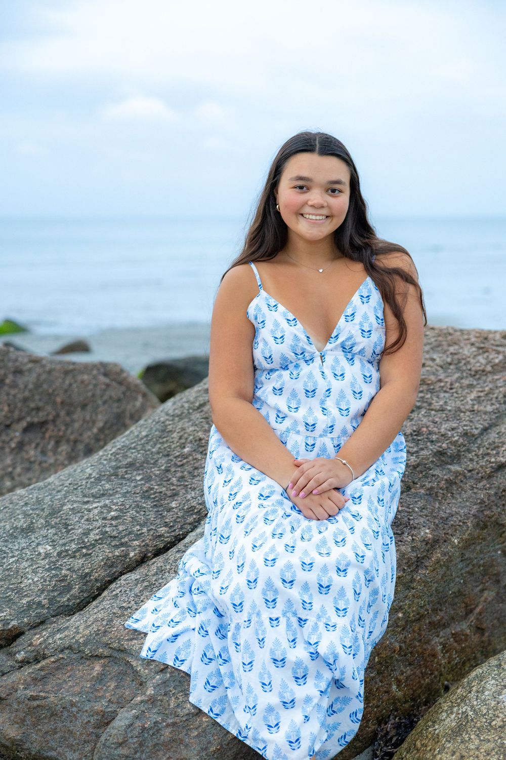 A woman in a blue and white butterfly print dress poses on coastal rocks against an ocean backdrop.