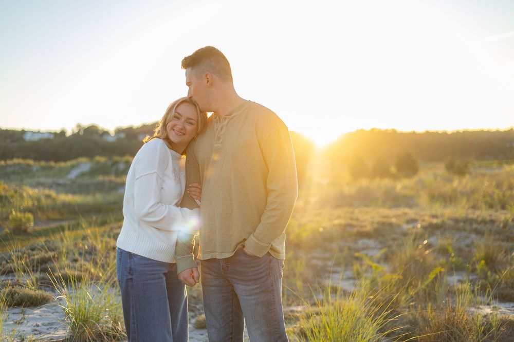 Couple embraces in golden sunlight on grassy hillside during sunset photoshoot.