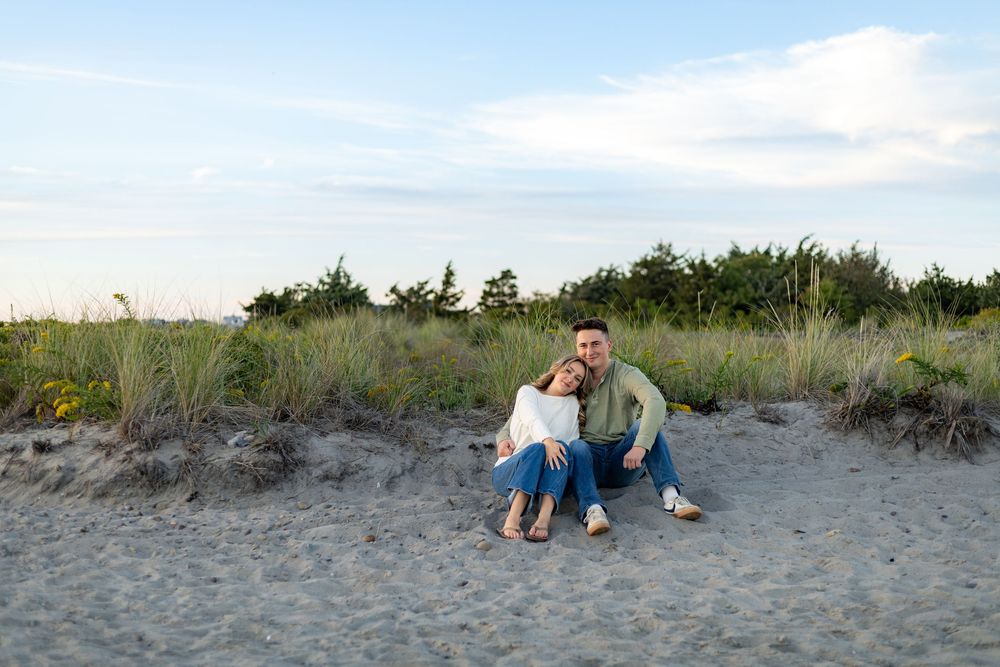People sitting together on a sandy beach with coastal vegetation and blue sky in the background.