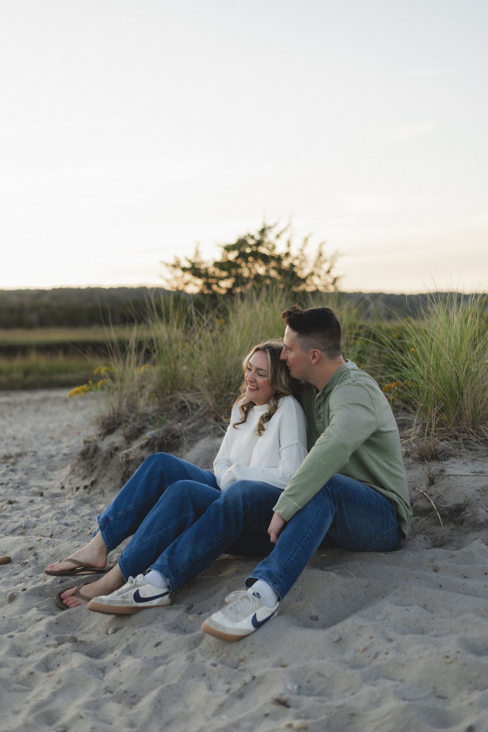 A couple relaxes together on a sandy beach at sunset wearing casual denim and sweaters.
