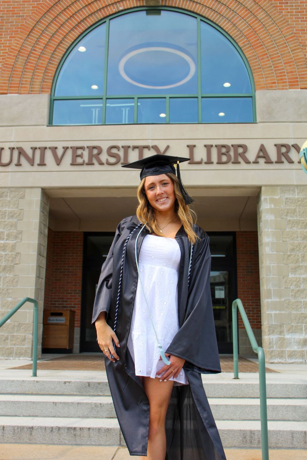 Graduate poses in front of University Library entrance wearing traditional graduation attire.