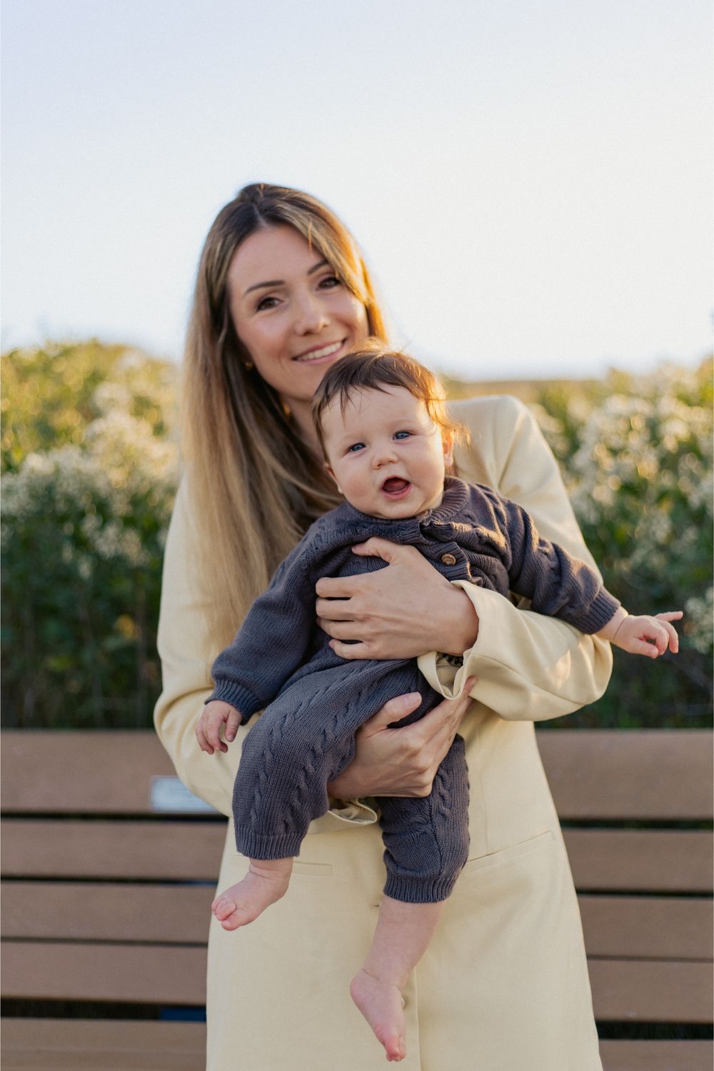 Parent holding a happy baby wearing navy overalls against a sunny outdoor background.
