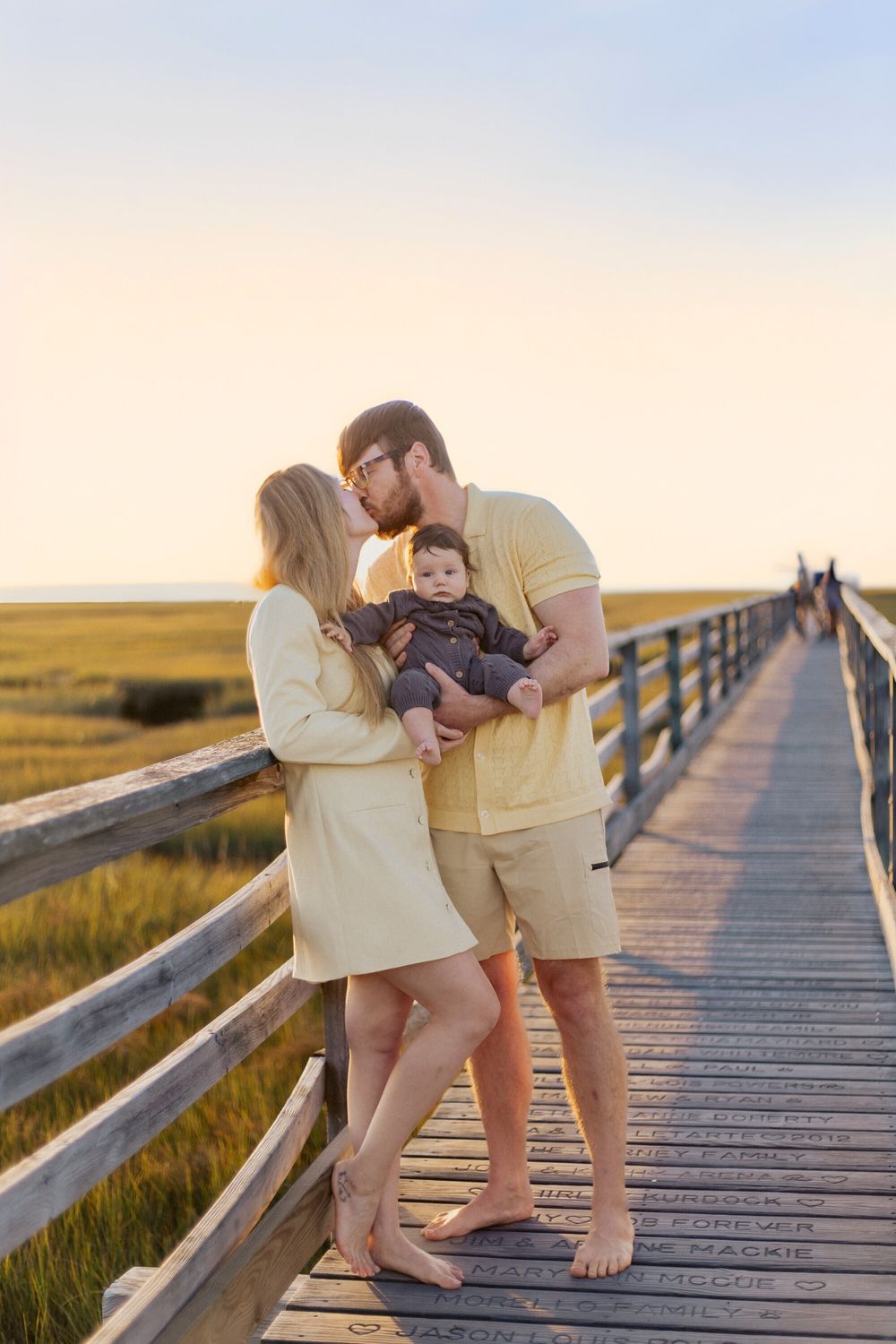 Family sharing a tender moment on a wooden boardwalk at sunset over coastal marshland.