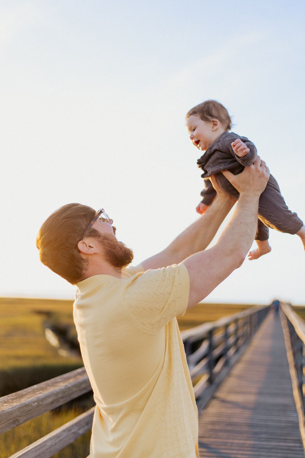A person in a yellow shirt lifts and plays with a small child on a wooden boardwalk at sunset.