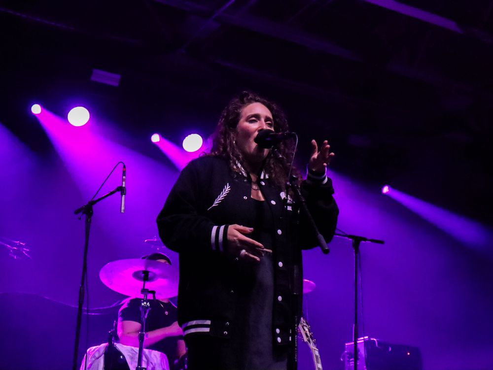 Concert performer singing on stage illuminated by purple and blue theatrical lighting.