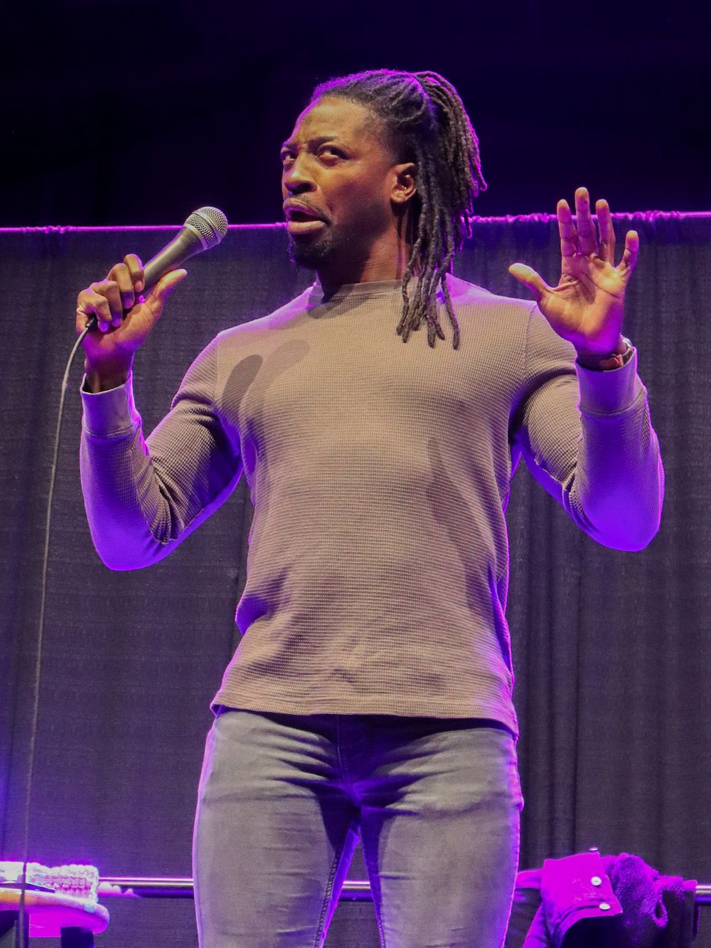 Stage performer in grey attire gesturing expressively during a performance under purple lighting.