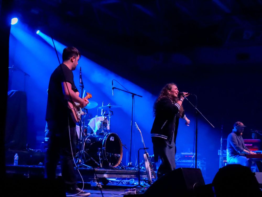 Band performing on stage with blue lighting and musical instruments including drums and guitars.