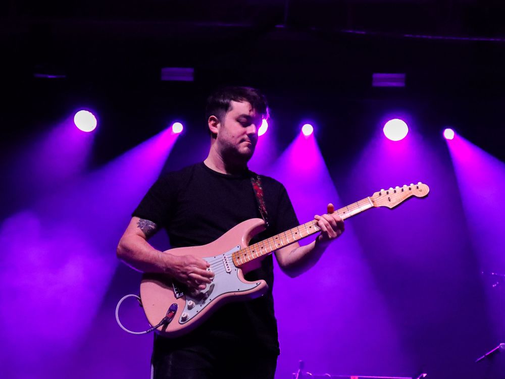 Guitarist playing electric guitar on stage bathed in purple concert lighting.