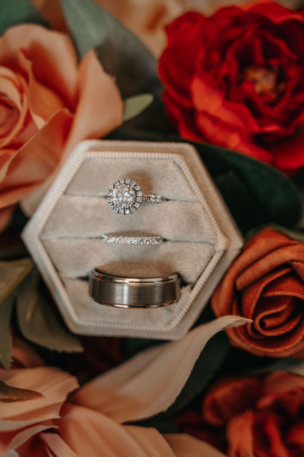 Diamond halo engagement ring, wedding band, and groom band displayed in velvet box with roses.