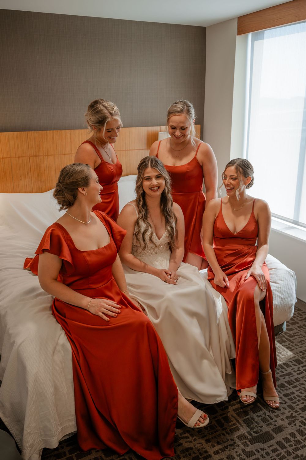 Bride in white gown sitting on hotel bed surrounded by four bridesmaids in rust satin dresses.