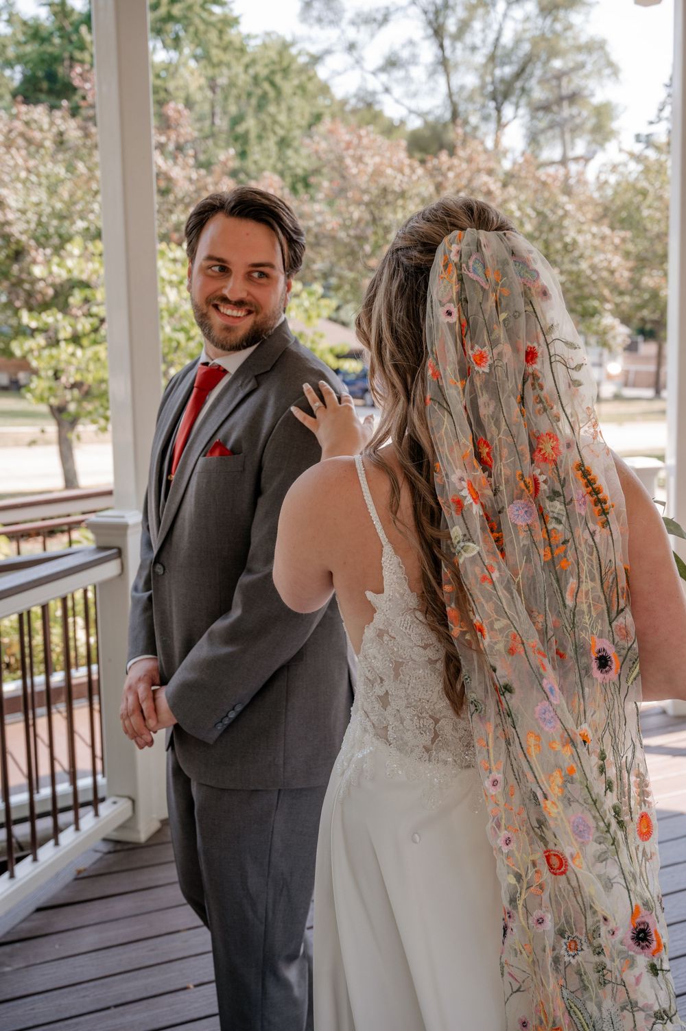 Groom in gray suit smiles at bride wearing floral embroidered veil during wedding first look on porch.