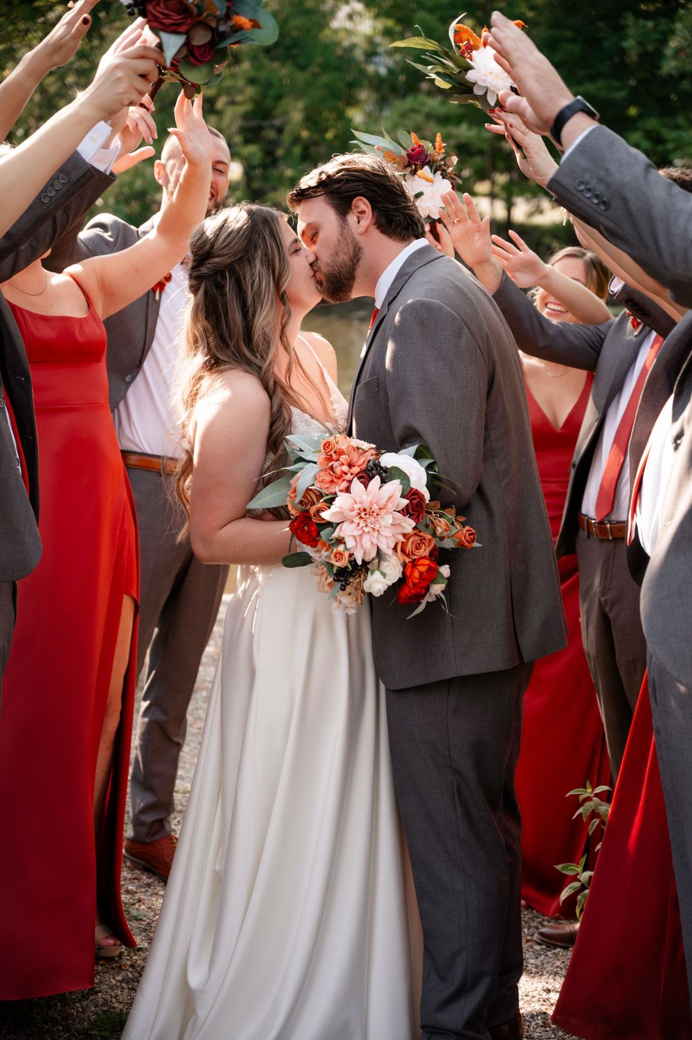 Bride and groom kiss surrounded by wedding party in red dresses and gray suits holding flowers.