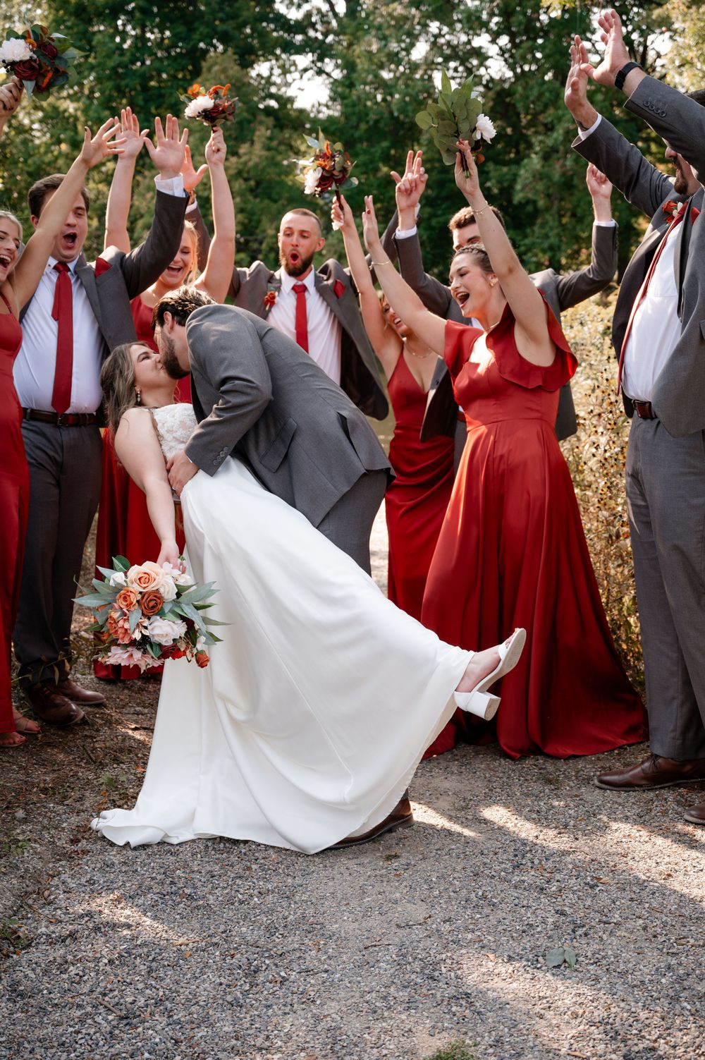 Groom dips bride for kiss as excited wedding party in red and gray celebrates outdoors.
