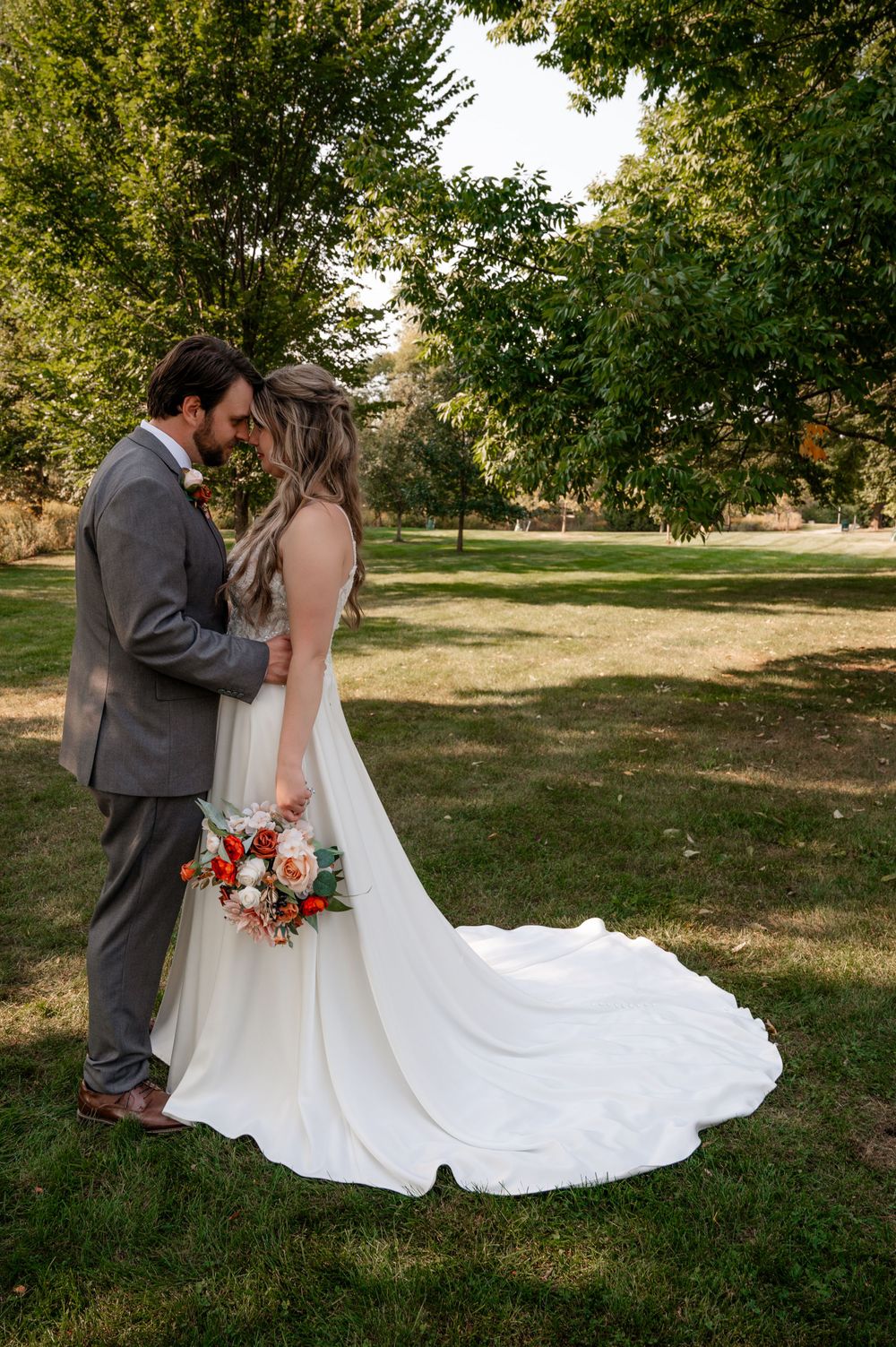 Bride and groom share intimate moment in park, bride holding orange and white floral bouquet.