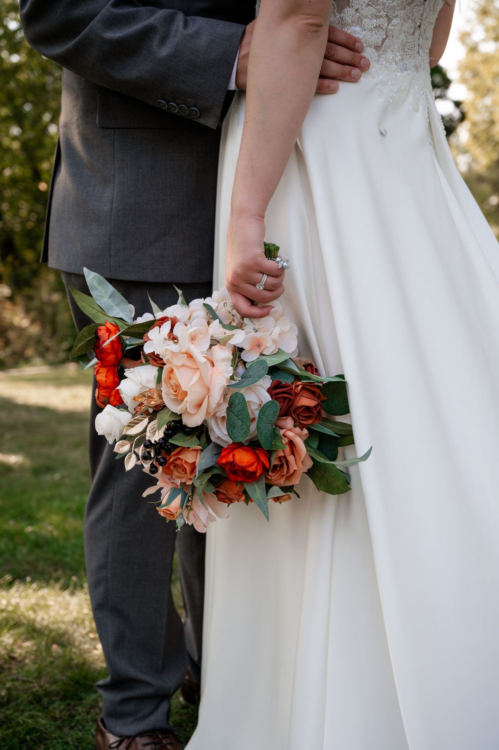 Bride holding a bouquet of red and blush peonies in a white gown beside groom in gray suit.