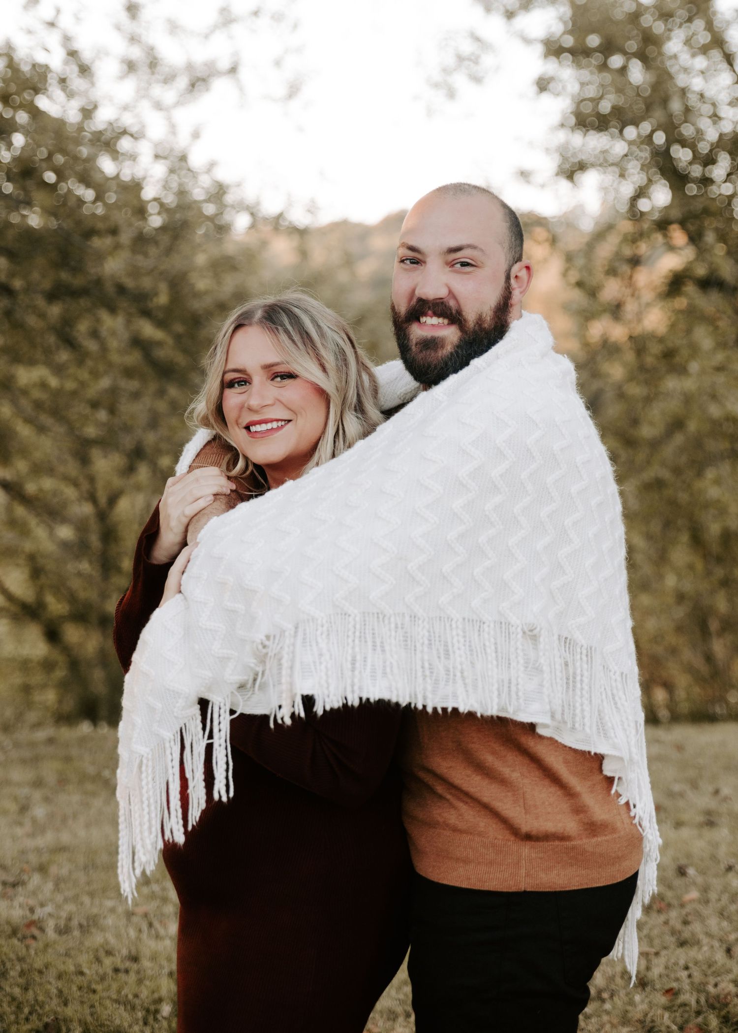 Playful couple wrapped in a white fringed blanket laughing in an autumn setting.