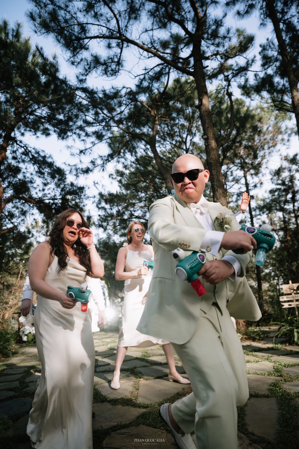 Groom in cream suit dances playfully with bridesmaids holding confetti poppers outdoors at a wedding.